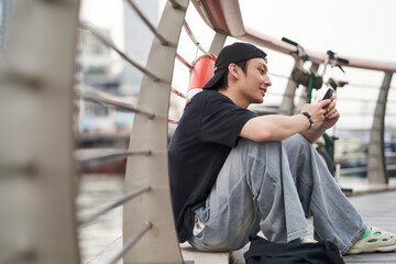 young Asian man sitting on ground chatting on smartphone at waterfront