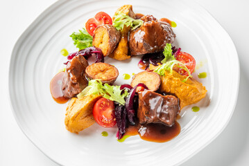 A gourmet dish featuring tender beef pieces with a rich brown sauce, roasted potatoes, cherry tomatoes, leafy greens, and a vibrant orange puree, artistically plated on a white dish.