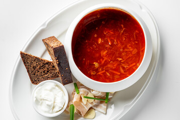 A bowl of traditional borscht with rye bread, sour cream, lard, and fresh garnishes on a white plate, showcasing Eastern European cuisine.