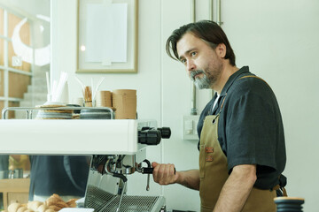 Obraz premium Senior Caucasian male barista wearing brown apron working attentively at espresso machine inside cozy small cafe preparing coffee drink with focus and craftsmanship., Concept small business