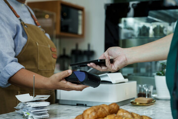Asian senior female barista adult receiving mobile payment from customer using smartphone at cafe demonstrating modern cashless transaction in cozy local family coffee shop