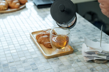 Closeup of tea being poured into double-wall glass next to croissant on wooden tray at cafe counter representing cozy beverage service in small local coffee shop