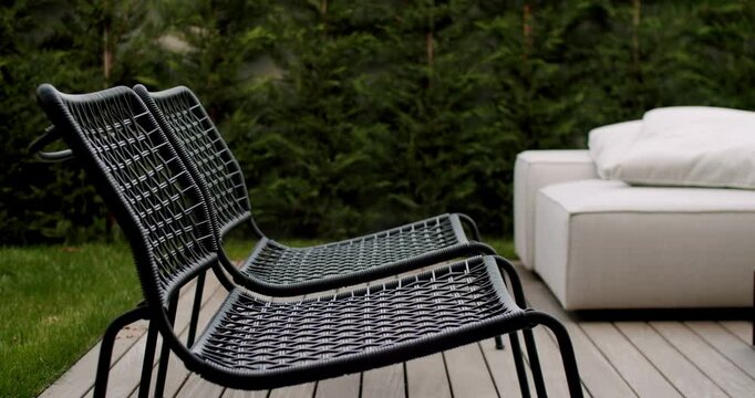 Two empty black wicker chairs stand on a wooden deck in a modern backyard. A white outdoor sofa is visible in the background. Lush green grass and manicured hedges create a serene atmosphere.