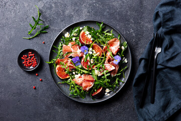 Arugula salad with fresh figs and prosciutto, jamon and edible flowers in a plate. Grey background....