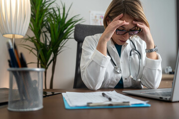 Stressed mature female doctor looking at laptop reading bad news online worried of mistake at workplace. Unhappy professional physician in tension feeling tired solving computer problem at work.