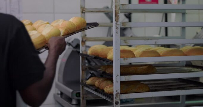 African American bakery worker in apron placing fresh baked bread on metal rack inside commercial kitchen