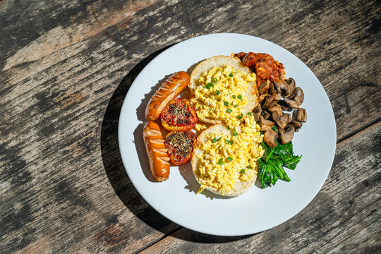 Big breakfast with bread toast, scrambled egg, sausages, roasted tomato, baked bean, sauted mushroom, and wilted spinach on white plate. Isolated by wooden table. Food Photography