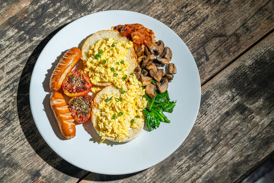 Big breakfast with bread toast, scrambled egg, sausages, roasted tomato, baked bean, sauted mushroom, and wilted spinach on white plate. Isolated by wooden table. Food Photography