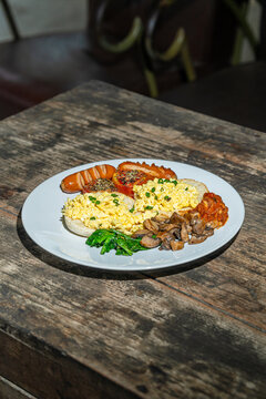 Big breakfast with bread toast, scrambled egg, sausages, roasted tomato, baked bean, sauted mushroom, and wilted spinach on white plate. Isolated by wooden table. Food Photography