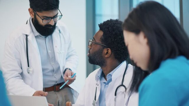 Indian and African American male doctors discussing medical topic, Asian female colleague reviewing notes beside them. Teamwork and exchange of ideas during clinical meeting.