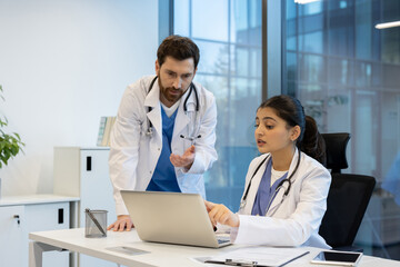 Two doctors collaborate in a modern office setting, focusing on a laptop while discussing medical information and data. The scene portrays professional teamwork.