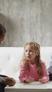 Cute little girl student tells new words to African-American teacher while brother sits nearby on sofa at home lesson with native speaker slow motion