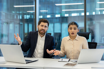 A shocked business team appears to be dealing with an unexpected issue while working at their office desk. They're using laptops.