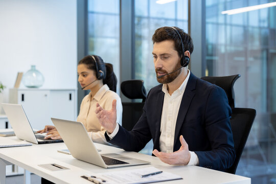 Two business professionals wearing headsets in an office setting, working on laptops with serious expressions. They appear to be customer service agents or call center representatives.