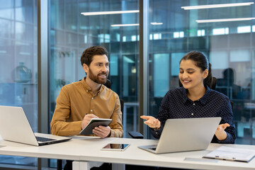 Two colleagues collaborate at a modern office desk, discussing a project on a laptop and taking notes on a tablet.