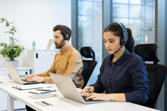 Two customer service representatives are working diligently at their computers, wearing headsets, in a modern office environment.