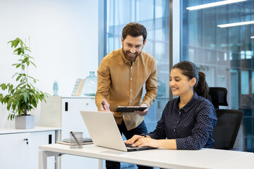 A cheerful colleague assists a female coworker as she works on a laptop in a bright modern office...
