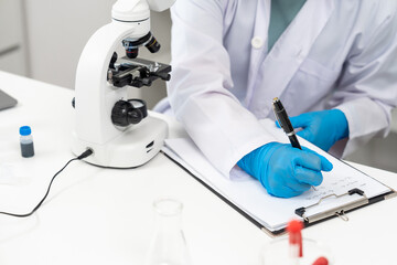 Closeup of asian scientist hand wearing blue glove writing research data on clipboard next to...