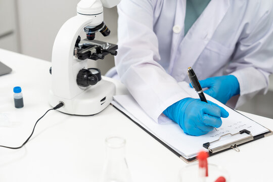 Closeup of asian scientist hand wearing blue glove writing research data on clipboard next to microscope in white laboratory, documenting analysis results during scientific experiment and observation