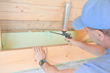 A man uses a caulking gun to apply insulation along a wooden beam in a home renovation project, highlighting the importance of energy efficiency and construction skills