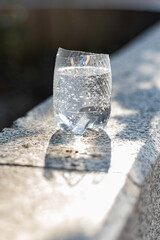 A cut plastic bottle filled with sparkling water sits on a stone surface in sunlight.
