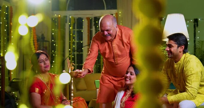 Lighting samai diya indoors during Diwali, Indian family of four including senior mother in saree, father, young girl, and husband smiling together celebrating Hindu festival with joy and tradition