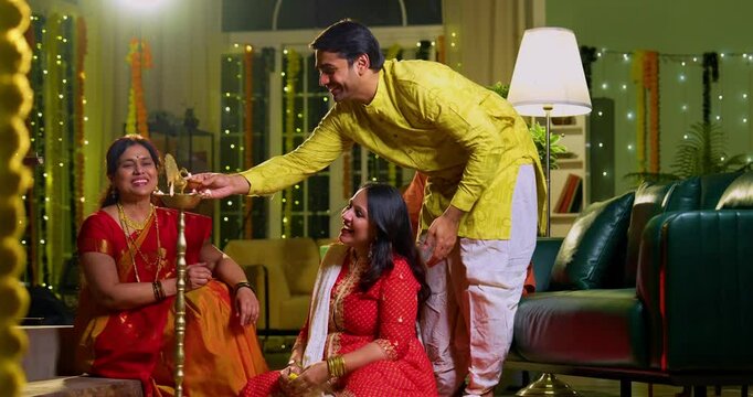Lighting samai diya indoors during Diwali, Indian family of four including senior mother in saree, father, young girl, and husband smiling together celebrating Hindu festival with joy and tradition