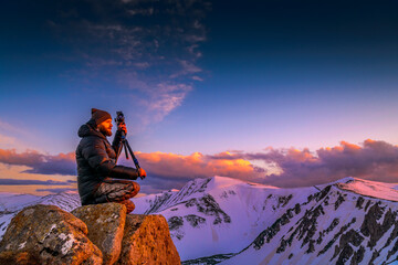 Male photographer in insulated winter gear squatting on rock, shooting snowy mountain ridge at vibrant sunset with medium format camera on tripod. © XArt