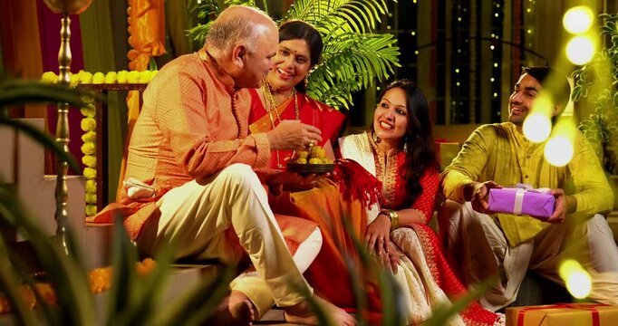 Holding laddus and smiling together, Indian family with young girl, senior mother in saree, father and husband in traditional wear celebrates Diwali indoors with diya, flower rangoli, and laughter