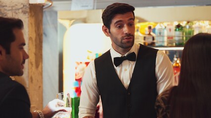 Smart smiling bartender preparing drinks putting ice into glass at luxurious bar while engaging lively conversation with VIP customer at vibrant warm lighting atmosphere pub and restaurant. Vinosity.