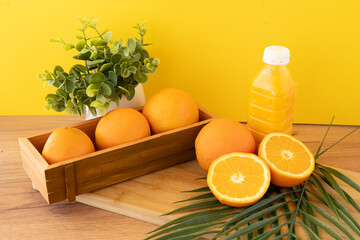 Fresh oranges and juice bottle on wood table with green plant and palm leaf in front of yellow backdrop Juice bottle and vibrant oranges with green leaves on wood table and bold yellow background back