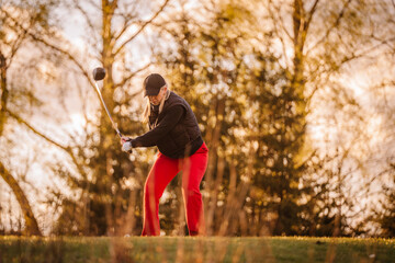 A woman golfer in red pants swings a club at sunset on a green course, framed by trees in soft golden light, mid-action moment..
