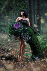 Slender beautiful brunette in the image of a forest fairy in full growth. Decorative cabbage instead of a bouquet. Artistic creative photography.