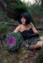 Slender brunette beauty in the image of a forest fairy sits on the ground. Decorative cabbage instead of a bouquet. Artistic creative photography.