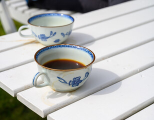Two beautifully designed cups holding dark coffee sit on a white wooden table in a bright garden. The sunlight enhances the moment, creating a tranquil atmosphere