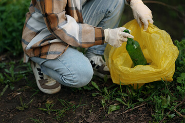 Man wearing gloves crouching and picking up litter in a forest or park, placing a glass bottle into...