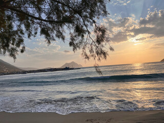 Sunset view of Aegiali harbor on Amorgos island, Greece, framed by tree branches with gentle waves...