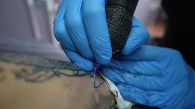 Close-up view of a tattoo artist in blue gloves creating a floral tattoo on a clients forearm. The tattoo machine is in motion as the artist outlines the design. Studio environment