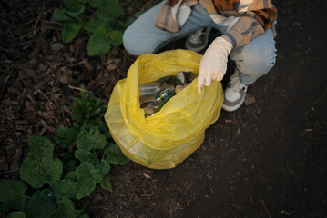 Top view of a yellow trash bag full of litter, held by a gloved hand. Person crouching beside the...