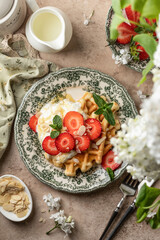 Homemade belgian waffles with strawberries, whipped cream and honey. Served on a rustic plate with spring flowers, top view