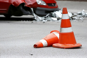 Traffic cones near a red car wreck, one standing and one fallen on a gray asphalt road with shattered glass. Safety measures at an accident scene.
