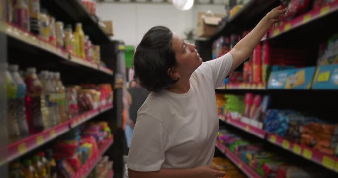 Latina woman fully extending arm to grab item from high shelf while shopping in grocery store aisle filled with snack products and drinks
