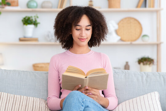 Spare time, hobby and rest and relax at home during covid-19 quarantine. Attractive smart millennial african american woman sitting on sofa and reading book in living room interior, free space
