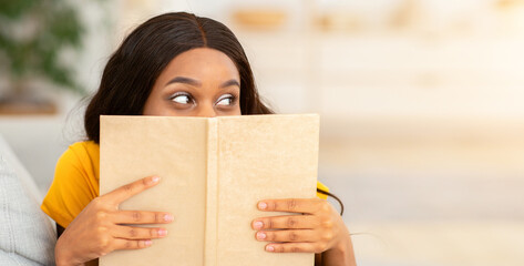 Stay home pastimes concept. Charming African American lady hiding behind interesting book on couch...