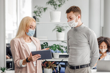 Obraz premium New normal, social distancing and boss with employee communication. Business woman in protective mask shows tablet to man, near workplace with laptop and antiseptic in interior of modern office, empty