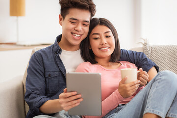 Fototapeta premium Asian Boyfriend And Girlfriend Using Tablet Computer Watching Movie Online Embracing Sitting On Sofa At Home Relaxing On Weekend Morning. Modern Gadgets, Chinese Family Lifestyle