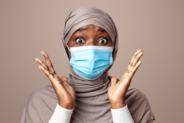 Coronavirus Panic. Studio portrait of black shocked muslim woman in protective medical face mask, holding hands near face and looking at camera, islamic lady posing in hijab over light background