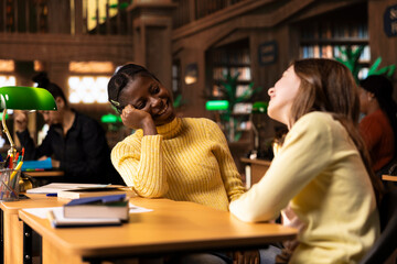 Cute teens friends gossiping together and laughing in the library, taking a break from school work and enjoying team project with each other. Young girls enjoying a cheerful conversation.