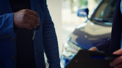 Showroom dealer completes paperwork after selling vehicle, shaking hands with buyer and offering car keys. Salesman in dealership signing contract with man, reaching agreement, camera A close up