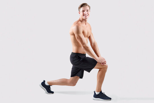 Shirtless millennial guy with bare torso doing lunges on light studio background, full length. Happy young sportsman with sexy body stretching his legs, working out, promoting healthy living